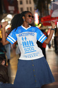 Woman wearing a blue and white t-shirt with text and graphics, standing in an urban setting with blurred background